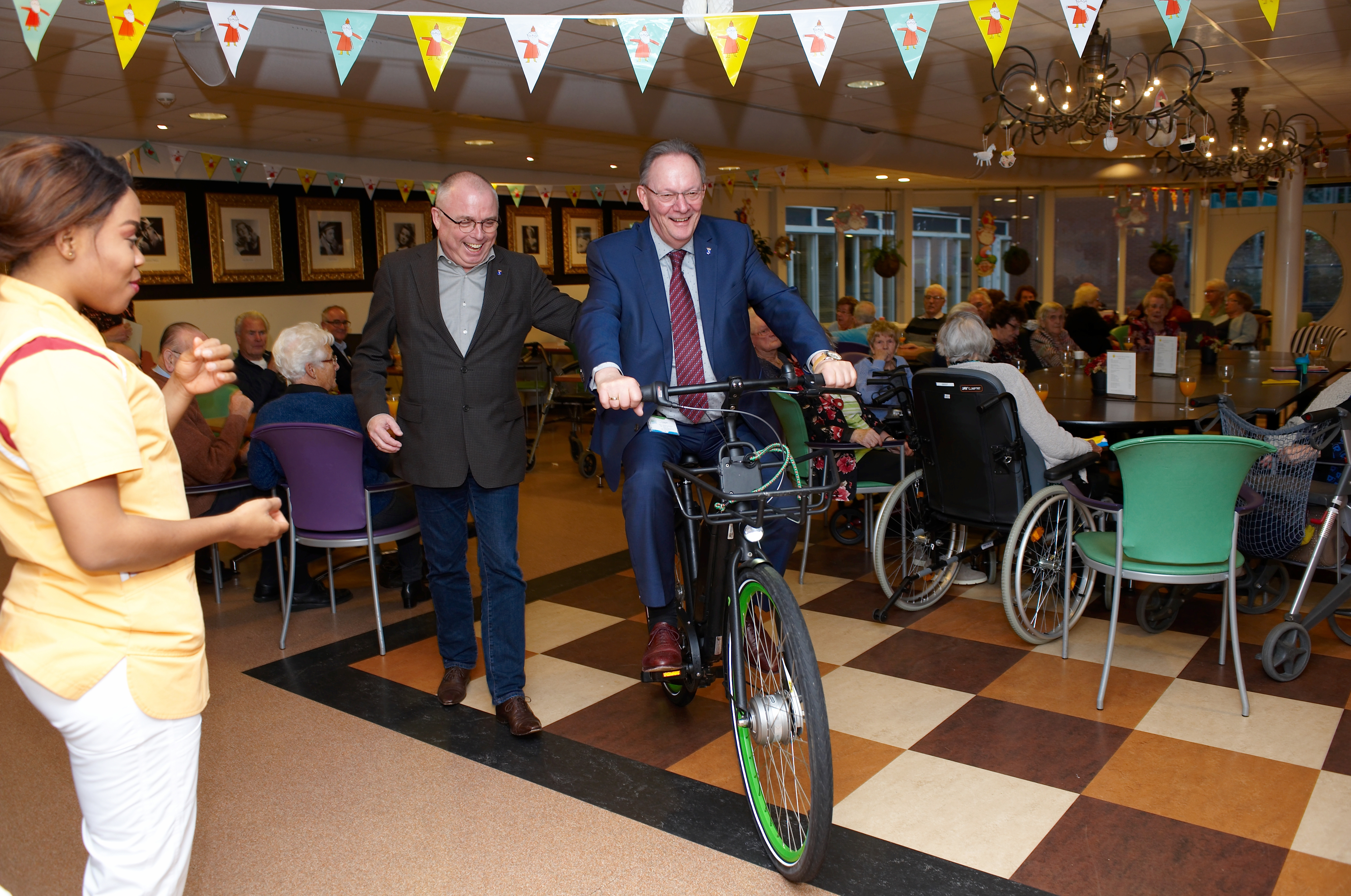 Burgemeester Peter Tange (op fiets) en wethouder Schalkwijk van Wormerland rijden een rondje door de zaal op een van de e-bikes van de Zorgcirkel Thuiszorg. (Foto: Goedheer Fotografie)