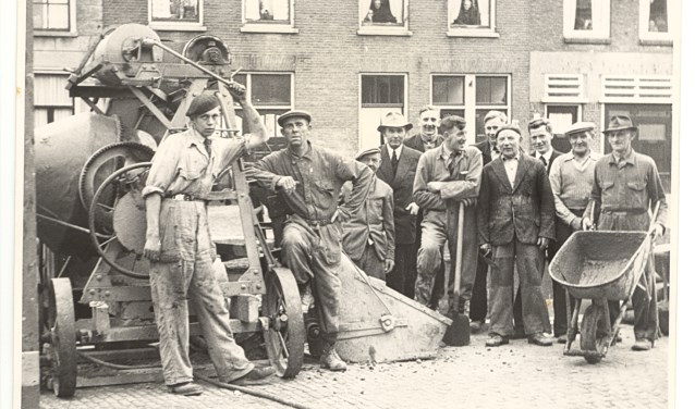 Bijschrift: Een fraaie foto van het personeel van Mensert aan het Achterom in 1955, toen de mannen nog van staal waren en mensen uit de ramen hingen als een foto werd gemaakt.