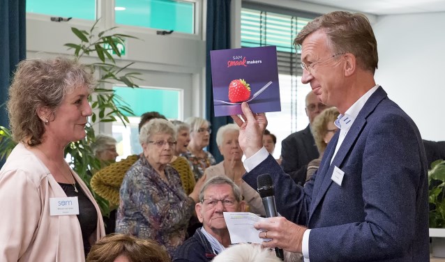 Ron de Haas, directeur Mozaïek Wonen, ontvangt uit handen van Mirjam van Veen, directeur SAM, het eerste exemplaar van het kookboekje dat de medewerkers van SAM hebben samengesteld