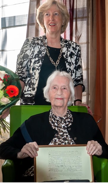 Mevrouw Timmers met haar ingelijste geboortecertificaat. Achter haar staat burgemeester Driessen, die de eeuwelinge namens de gemeente Leiderdorp kwam feliciteren.